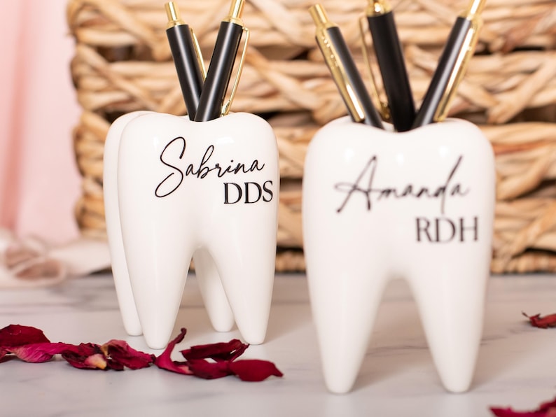 May include: Two white ceramic tooth-shaped pencil holders. The holder on the left has the name Sabrina DDS written on it. The holder on the right has the name Amanda RDH written on it.