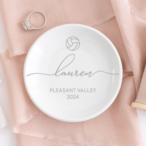 May include: White ceramic ring dish with a volleyball graphic, the name "Lauren", "Pleasant Valley", and the year "2024" printed in gray. A silver ring with a large, clear stone sits nearby.