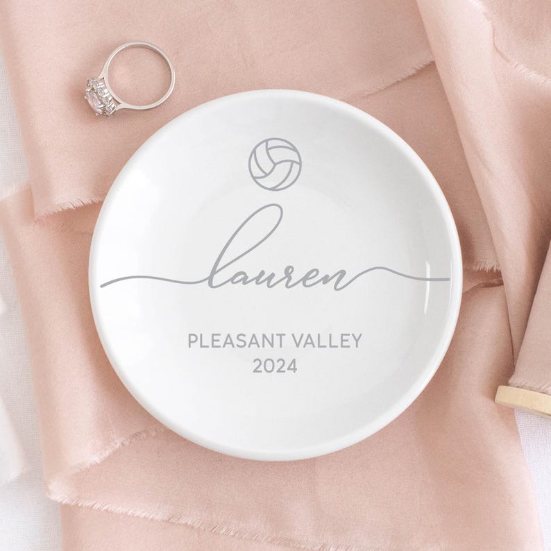 May include: White ceramic ring dish with a volleyball graphic, the name "Lauren", "Pleasant Valley", and the year "2024" printed in gray. A silver ring with a large, clear stone sits nearby.