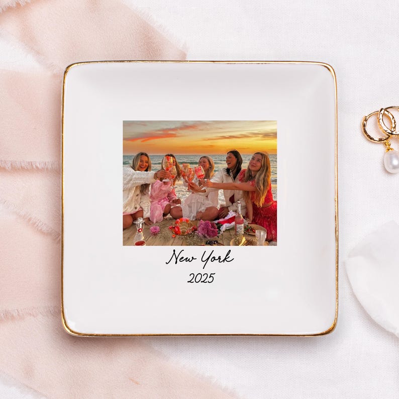 Puede incluir: Plato cuadrado de cer&aacute;mica blanca con borde dorado. El centro presenta una fotograf&iacute;a de un grupo de mujeres brindando en una playa al atardecer. Las palabras "New York" y "2025" est&aacute;n escritas debajo.