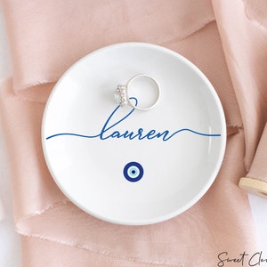 May include: White ceramic ring dish with a blue evil eye design and the name "Lauren" written in cursive.
