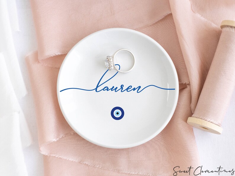May include: White ceramic ring dish with a blue evil eye design and the name "Lauren" written in cursive.