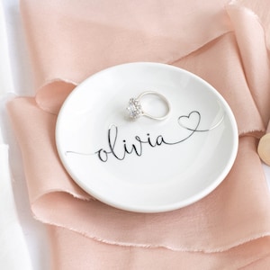 May include: A white ceramic ring dish with the name "olivia" written in cursive script and a small heart design. The dish is sitting on a light pink satin ribbon.
