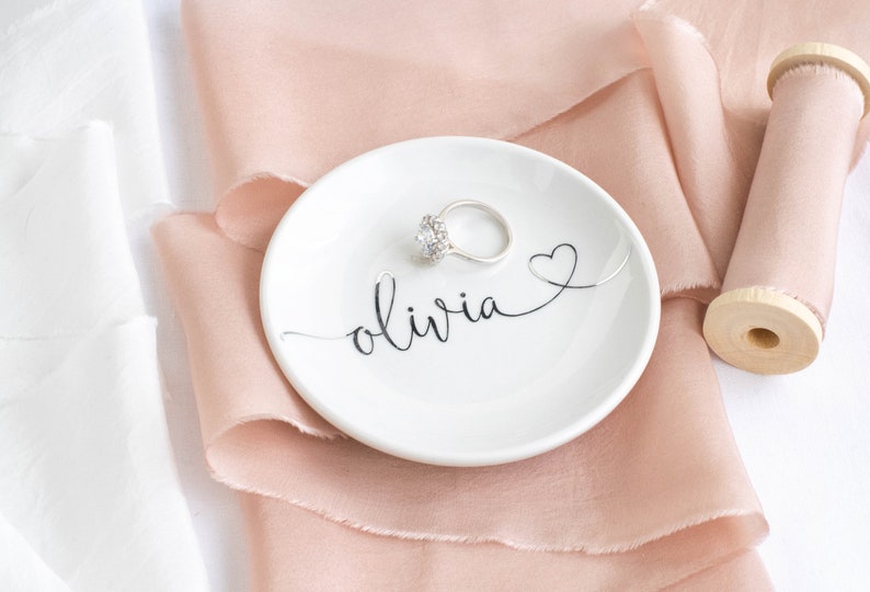 May include: A white ceramic ring dish with the name "olivia" written in cursive script and a small heart design. The dish is sitting on a light pink satin ribbon.