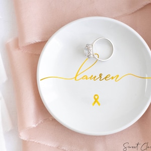 Op de afbeelding: Een witte keramische ringenschotel met een gouden lint en de naam "lauren" in cursief.