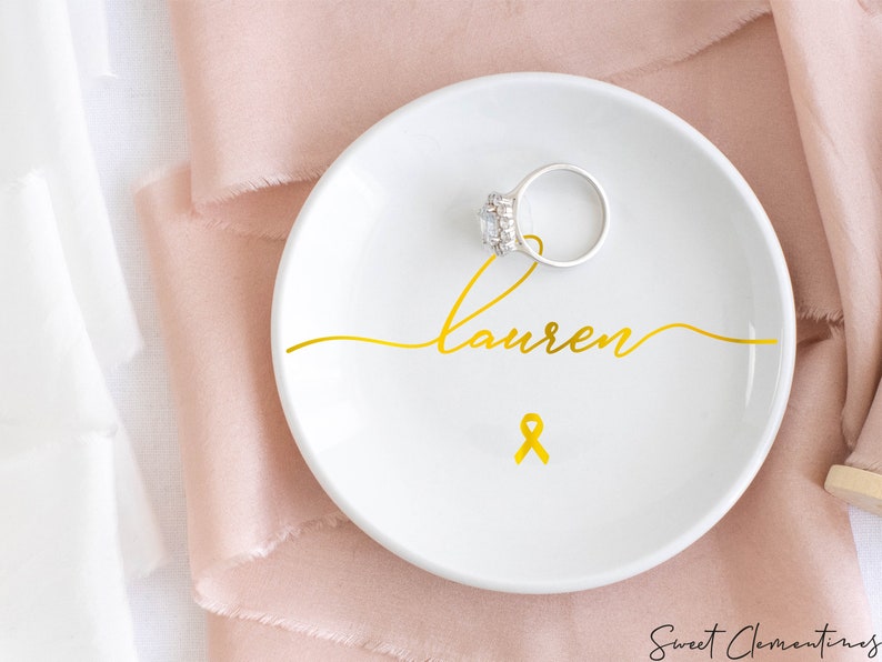 Op de afbeelding: Een witte keramische ringenschotel met een gouden lint en de naam "lauren" in cursief.