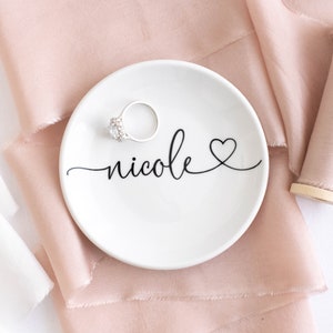 May include: White ceramic ring dish with the name "nicole" written in cursive with a small heart at the end. A silver ring with a clear stone is sitting on the dish.