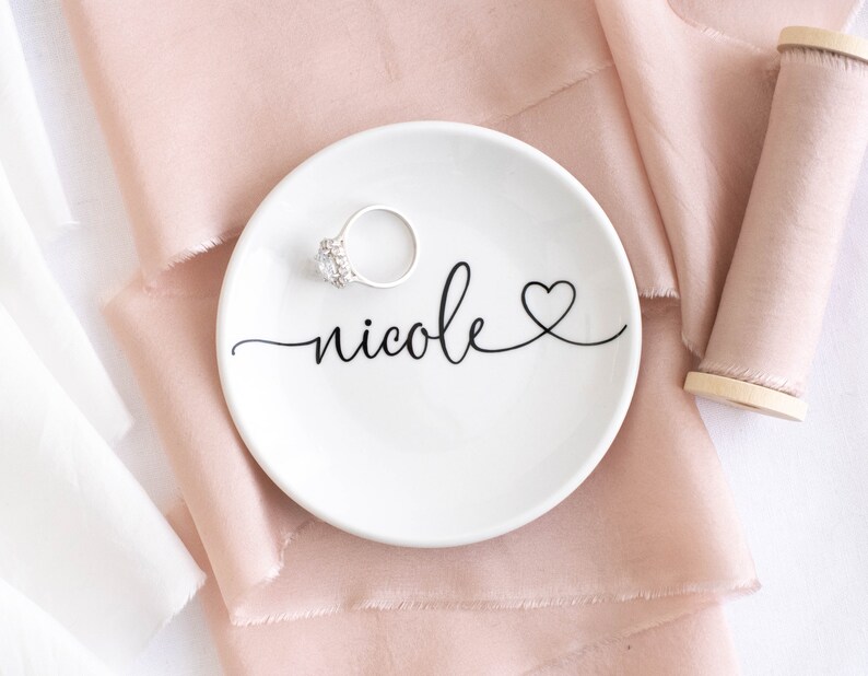 May include: White ceramic ring dish with the name "nicole" written in cursive with a small heart at the end. A silver ring with a clear stone is sitting on the dish.