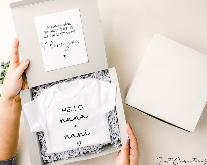 Op de afbeelding: Een witte romper met de tekst "HELLO nana + nani" in zwarte letters en een klein hartje. De romper zit in een witte cadeaudoos met wit tissuepapier. Een kaart met de tekst "HI NANA & NANI, WE HAVEN'T MET YET BUT I ALREADY KNOW... I love you" ligt bovenop de doos.