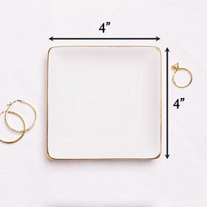 Puede incluir: Una bandeja cuadrada blanca para baratijas con borde dorado, de 10 cm por cada lado. Un anillo de oro con un diamante y un par de pendientes de aro dorados est&aacute;n junto a la bandeja. La bandeja est&aacute; dise&ntilde;ada para joyas.
