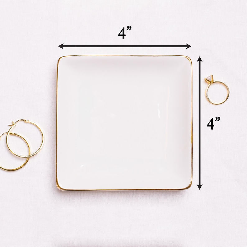 Puede incluir: Una bandeja cuadrada blanca para baratijas con borde dorado, de 10 cm por cada lado. Un anillo de oro con un diamante y un par de pendientes de aro dorados est&aacute;n junto a la bandeja. La bandeja est&aacute; dise&ntilde;ada para joyas.