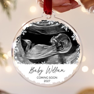 May include: A clear, circular acrylic ornament featuring a black and white ultrasound image of a fetus. The design includes white snowflake accents, the text Baby William, and the words COMING SOON 2027. A red ribbon is attached to the top loop.