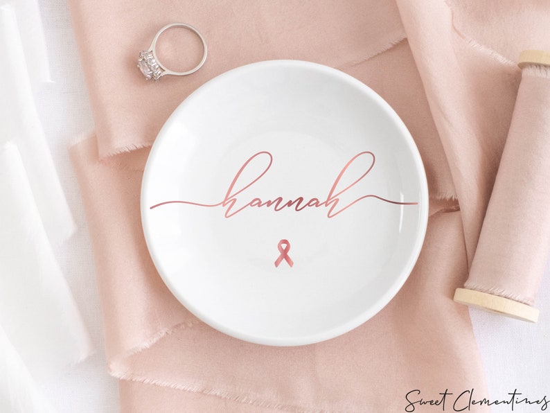 Op de afbeelding: Een witte keramische ringenschotel met de naam "Hannah" geschreven in ros&eacute;goud schrift. Een ros&eacute;gouden lint is gecentreerd onder de naam.