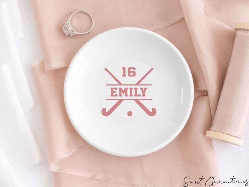May include: White ceramic ring dish with a pink design of two crossed field hockey sticks, the number 16, and the name "EMILY".