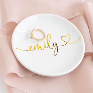 May include: White ceramic ring dish with gold lettering that says "emily" and a gold heart. A gold ring is sitting on the dish.