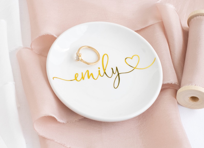 May include: White ceramic ring dish with gold lettering that says "emily" and a gold heart. A gold ring is sitting on the dish.