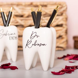 May include: Two white ceramic tooth-shaped pencil holders. The holder on the left has the text "Sabrina DDS" and the holder on the right has the text "Dr. Robinson".
