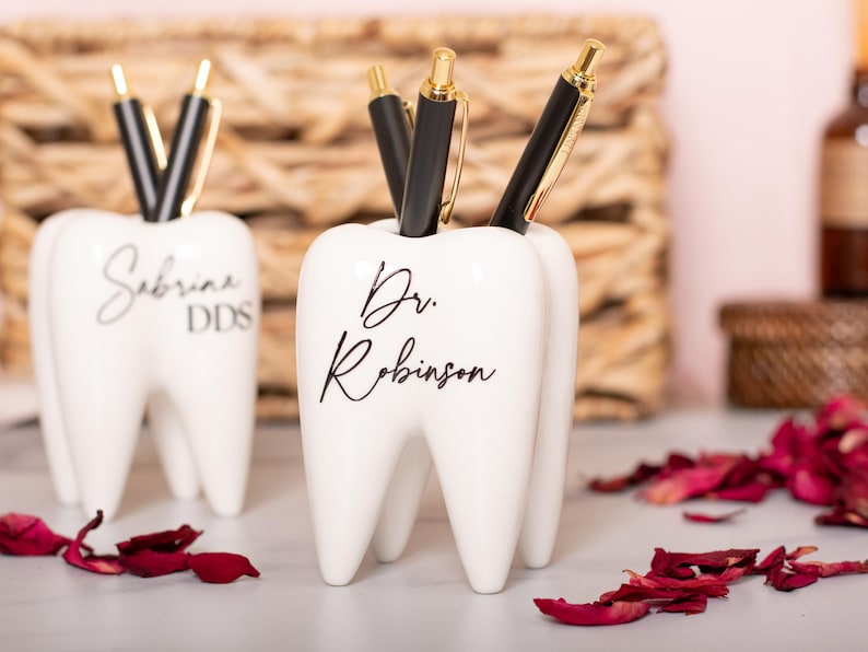 May include: Two white ceramic tooth-shaped pencil holders. The holder on the left has the text "Sabrina DDS" and the holder on the right has the text "Dr. Robinson".
