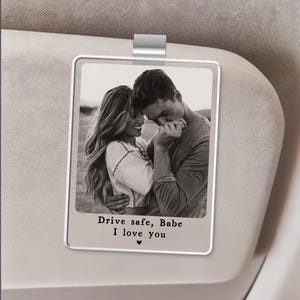 May include: A rectangular, clear car visor photo clip with a black and white image of a couple embracing. The text "Drive safe, Babe I love you" is printed below the image, with a small heart symbol.