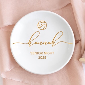 May include: A white ceramic ring dish with gold text and a volleyball graphic. The dish features the name "Hannah", the words "Senior Night", and the year "2025". The dish is round and sits on a pink fabric.