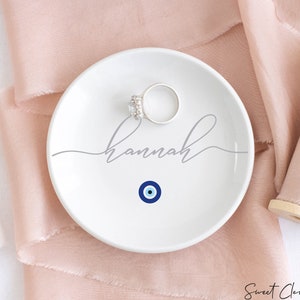 May include: White ceramic ring dish with a blue evil eye design and the name "Hannah" written in gray cursive script.