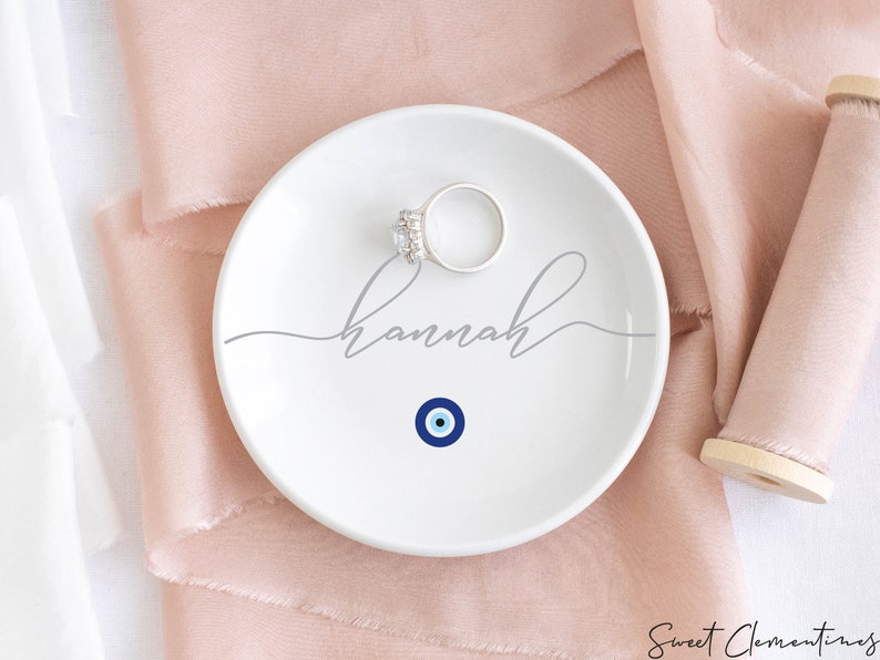 May include: White ceramic ring dish with a blue evil eye design and the name "Hannah" written in gray cursive script.