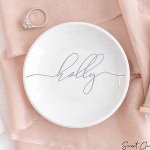 May include: A white ceramic ring dish with the word "holly" written in cursive on it. The dish is sitting on a pink fabric background.