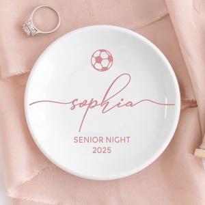 Pode incluir: Uma tigela de cerâmica branca para anéis com um desenho de bola de futebol rosa e o nome "Sophia" em escrita cursiva. As palavras "Senior Night 2025" também estão impressas. Um anel de prata é visível.