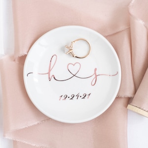 May include: White ceramic ring dish with a rose gold inscription that reads "h&j" with a heart symbol and the date "12.24.21". A gold ring with a clear stone is sitting on the dish.