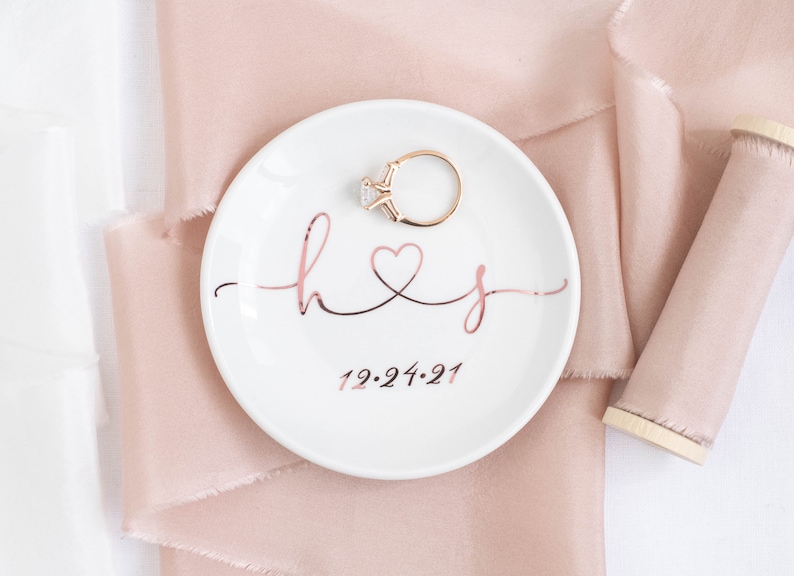 May include: White ceramic ring dish with a rose gold inscription that reads "h&j" with a heart symbol and the date "12.24.21". A gold ring with a clear stone is sitting on the dish.