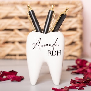 May include: White ceramic tooth-shaped desk organizer with the name "Amanda RDH" written on it. Three black pens are in the organizer.