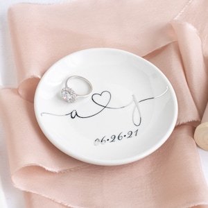 May include: White ceramic ring dish with a black cursive design of the letters "A" and "J" intertwined with a heart. The date "06.26.21" is printed below the letters. A silver ring with a clear stone is sitting on the dish.