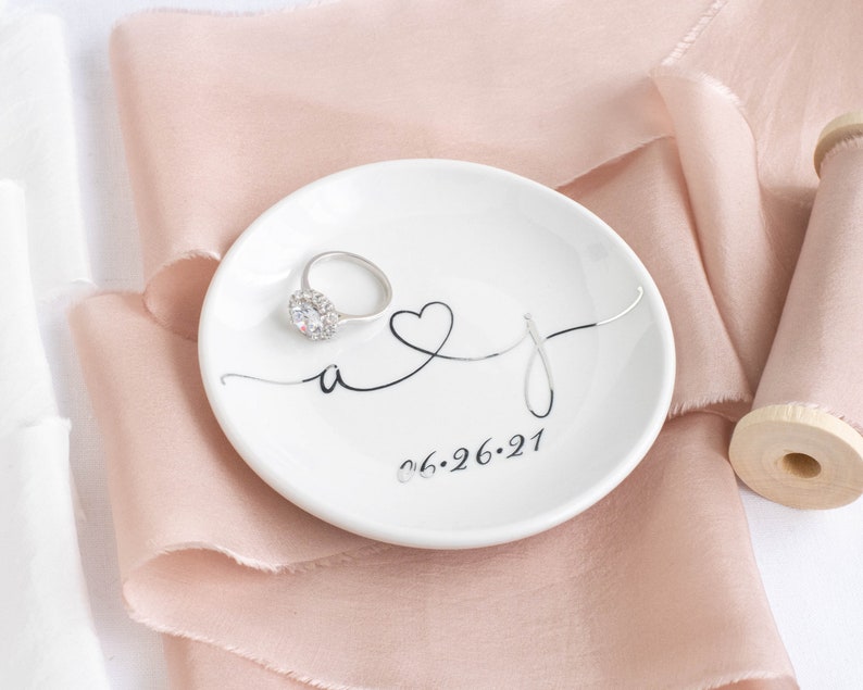 May include: White ceramic ring dish with a black cursive design of the letters "A" and "J" intertwined with a heart. The date "06.26.21" is printed below the letters. A silver ring with a clear stone is sitting on the dish.