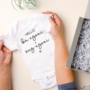 May include: A white baby onesie with the words "HELLO b&agrave; ngoại + &ocirc;ng ngoại" in black script. The onesie is held by hands, next to a gray gift box filled with white shredded paper.