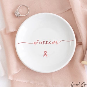 Op de afbeelding: Een witte keramische ringenschotel met het woord "warrior" geschreven in ros&eacute;goud schrift. Een ros&eacute;gouden lint is gecentreerd onder het woord.