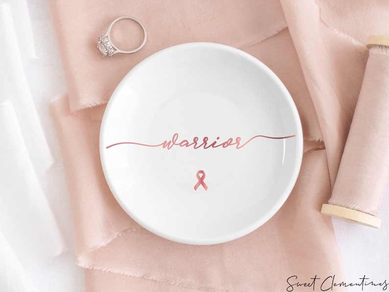 Op de afbeelding: Een witte keramische ringenschotel met het woord "warrior" geschreven in ros&eacute;goud schrift. Een ros&eacute;gouden lint is gecentreerd onder het woord.