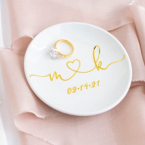 May include: White ceramic ring dish with gold lettering that says "m & k" with a heart between the letters and the date "02.14.21" below. A gold ring with a diamond is sitting on the dish.
