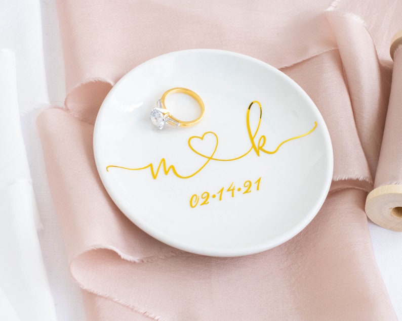 May include: White ceramic ring dish with gold lettering that says "m & k" with a heart between the letters and the date "02.14.21" below. A gold ring with a diamond is sitting on the dish.