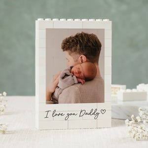 May include: White brick-style photo frame with a picture of a person holding a baby. The frame has the text "I love you Daddy" in black script. The frame is a unique gift for Father's Day or a new dad.