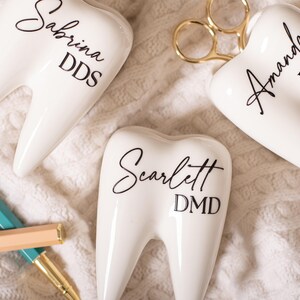 May include: Three white ceramic tooth-shaped holders with black lettering. The holders are labeled "Sabrina DDS", "Amanda RDH", and "Scarlett DMD".