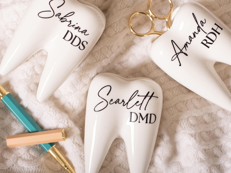May include: Three white ceramic tooth-shaped holders with black lettering. The holders are labeled "Sabrina DDS", "Amanda RDH", and "Scarlett DMD".