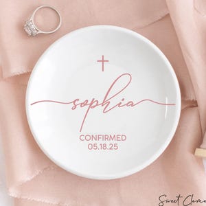 May include: White ceramic ring dish with a pink cross and the name "Sophia" written in cursive. The text "CONFIRMED 05.18.25" is also written on the dish.