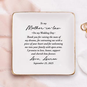 Peut inclure: Un vide-poches en céramique blanc avec un bord doré, portant un message sincère à une belle-mère. Le texte comprend une date de mariage et une signature. Boucles d'oreilles créoles dorées avec des perles.