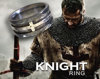 May include: A silver ring with a gold cross design. The ring is textured and has a wide band. The ring is shown on a model wearing chainmail armor and holding a sword. The text "KNIGHT RING" is visible below the ring.