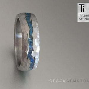 May include: A silver titanium ring with a hammered finish and a blue gemstone inlay. The ring is a wedding band style with a flat top and rounded edges. The text "CrackGemstone" is visible in the lower right corner of the image.