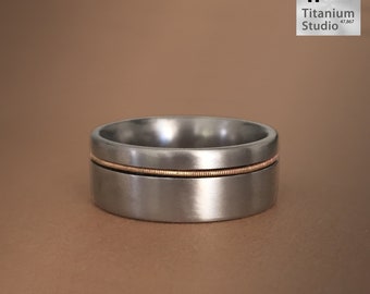 May include: A silver titanium ring with a thin gold guitar string inlay.  The ring is a brushed finish and has a beveled edge.  "Guitar String Ring" is written below the ring.