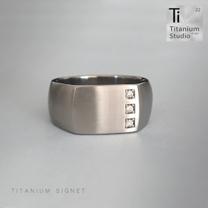 May include: A titanium signet ring with a brushed finish and three small diamonds set in a vertical line. The ring is a classic style and would make a great gift for a man.