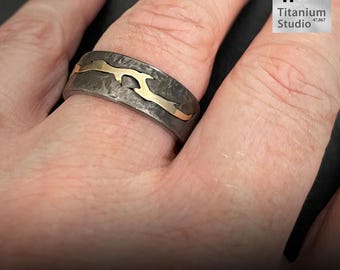 Puede incluir: Un anillo de titanio agrietado con una incrustación dorada. El anillo tiene una superficie texturizada de color gris oscuro y el texto "Cracked Titanium" en la parte inferior. El anillo es de Titanium Studio.