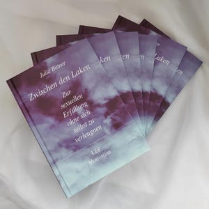 Könnte beinhalten: Ein Stapel identischer Bücher mit violett-weißem Wolkenmuster auf dem Einband. Der Text auf dem Cover lautet: Julia Bittner, Zwischen den Laken, Zur sexuellen Erfüllung ohne sich selbst zu verleugnen und LEP Motivation. Die Bücher sind fächerförmig angeordnet.