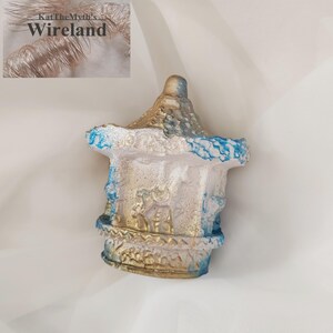 Op de afbeelding: Een decoratieve, miniatuur pagodesculptuur. De pagode is geschilderd in wit, goud en blauw, met een textuur. Het dak en de basis hebben gouden accenten en de randen zijn blauw omlijnd. De sculptuur staat op een witte achtergrond.